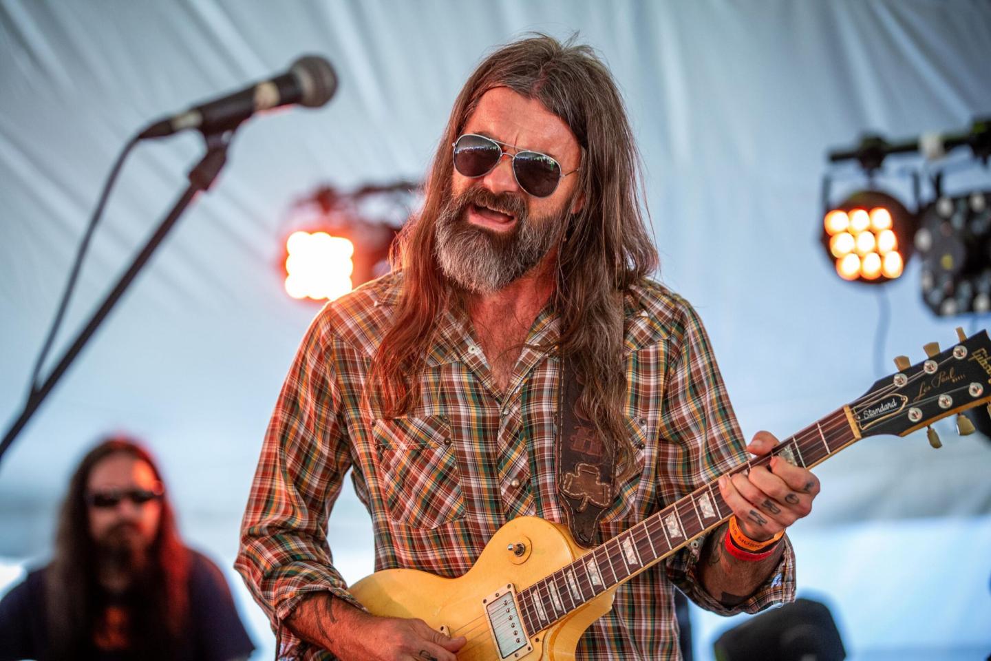 Musician in sunglasses playing guitar on stage under a tent.