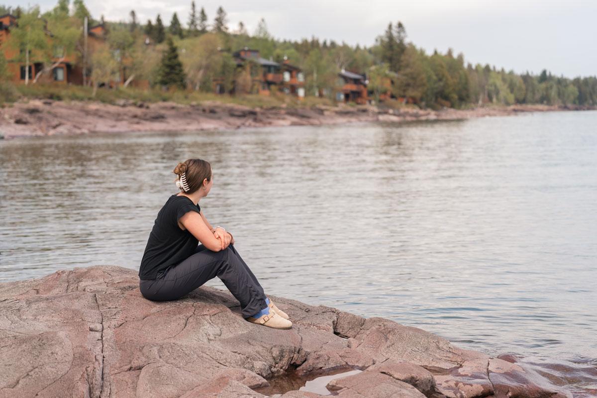 Person sitting on rocks by a lake, looking at distant cabins and trees.