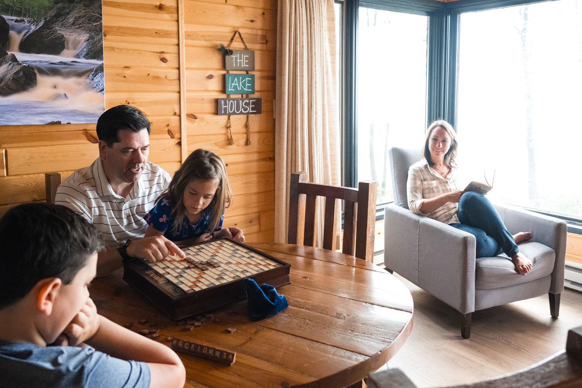 Family playing Scrabble at a wooden table, woman reading by window.