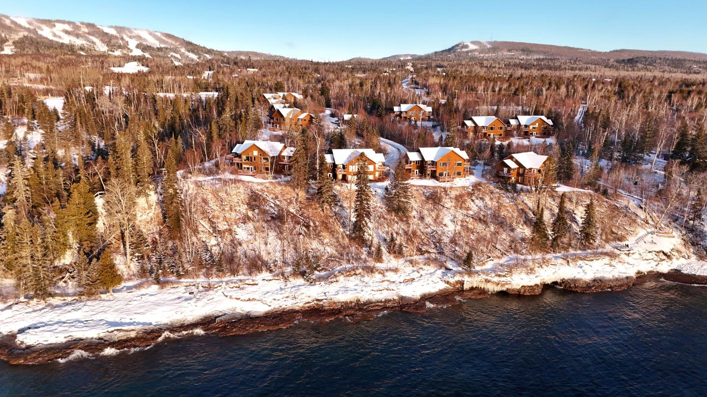 Snow-covered cabins on a forested hillside by a lake in winter.