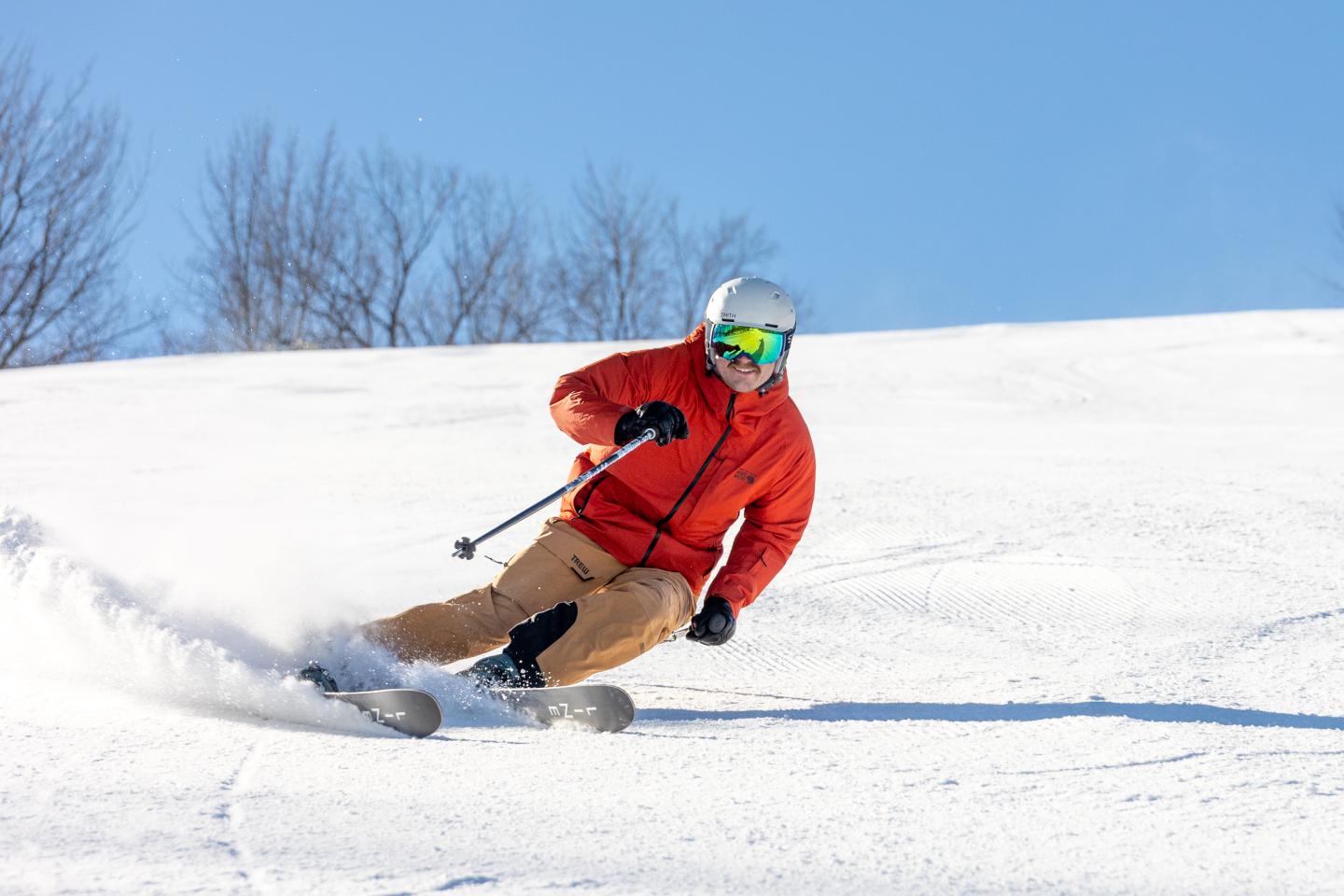 Skier in orange jacket carving down snowy slope under clear blue sky.