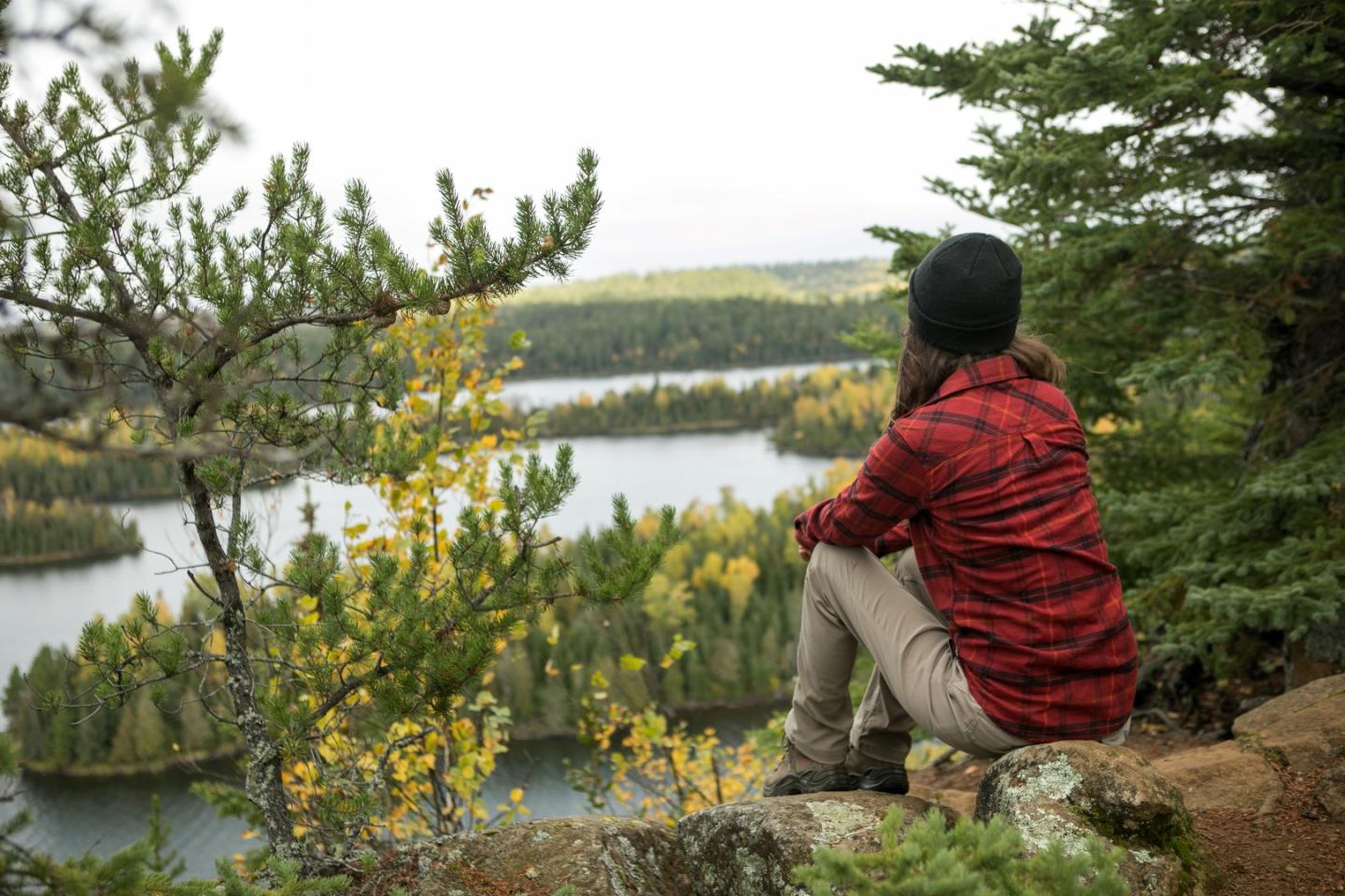 Person in red jacket sitting on rock, overlooking a forested lake.