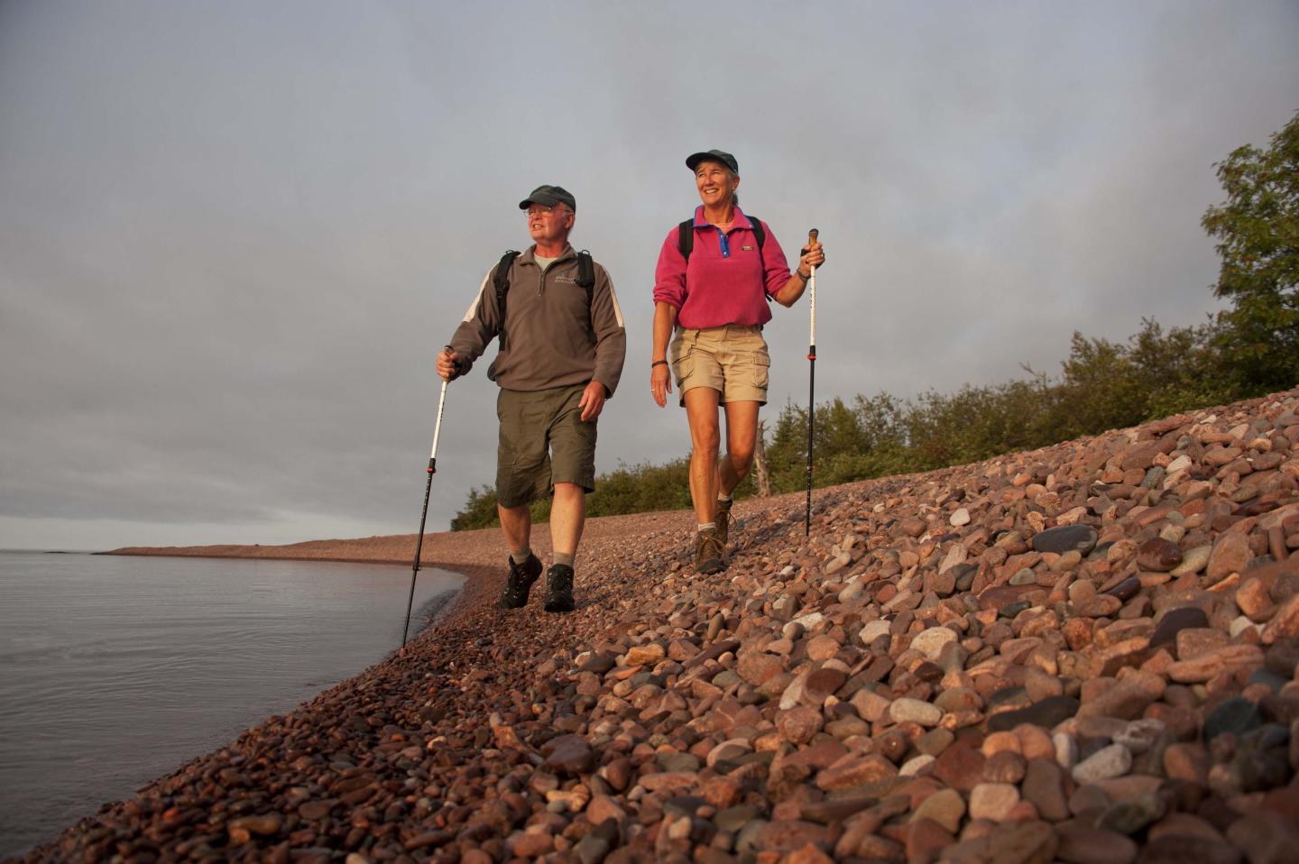 Two hikers with poles walk along a rocky lakeshore at sunset.