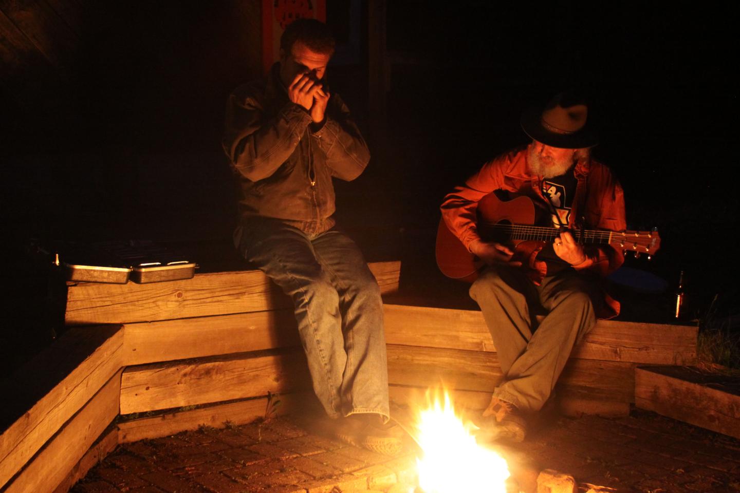 Two people by a campfire, one playing guitar, the other holding a harmonica.