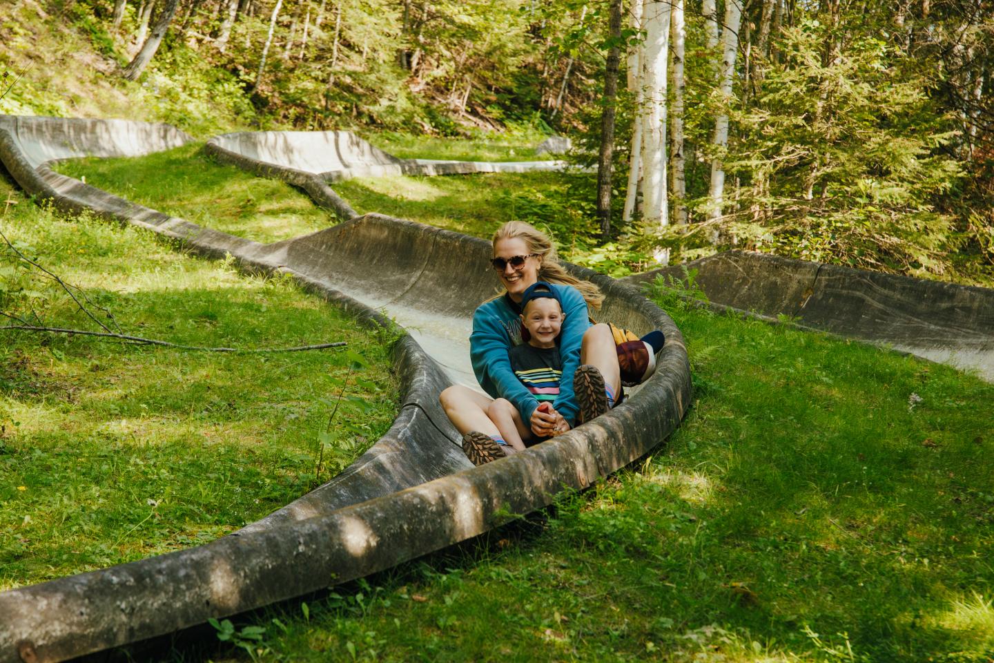 Two people enjoying a ride on a concrete slide in a forest, greenery surrounds them.