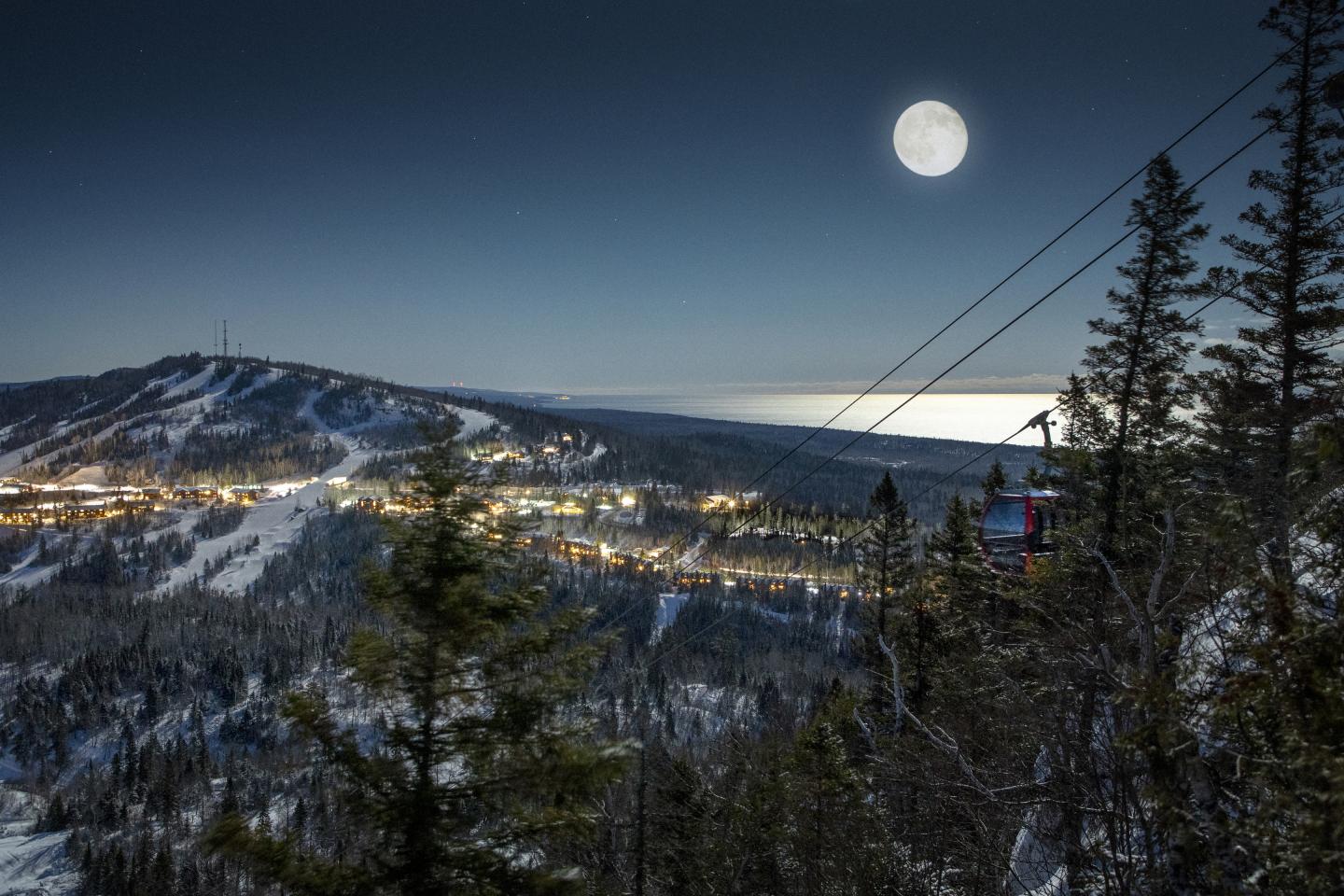 Snowy mountain landscape under a full moon, with distant city lights.