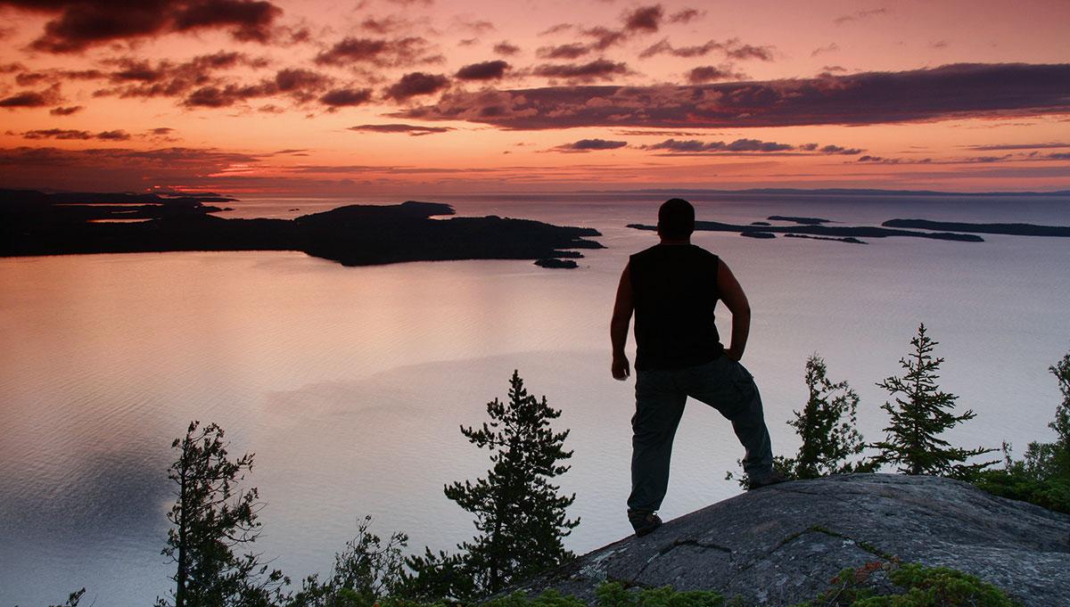 Silhouette of person on cliff overlooking lake at sunset.