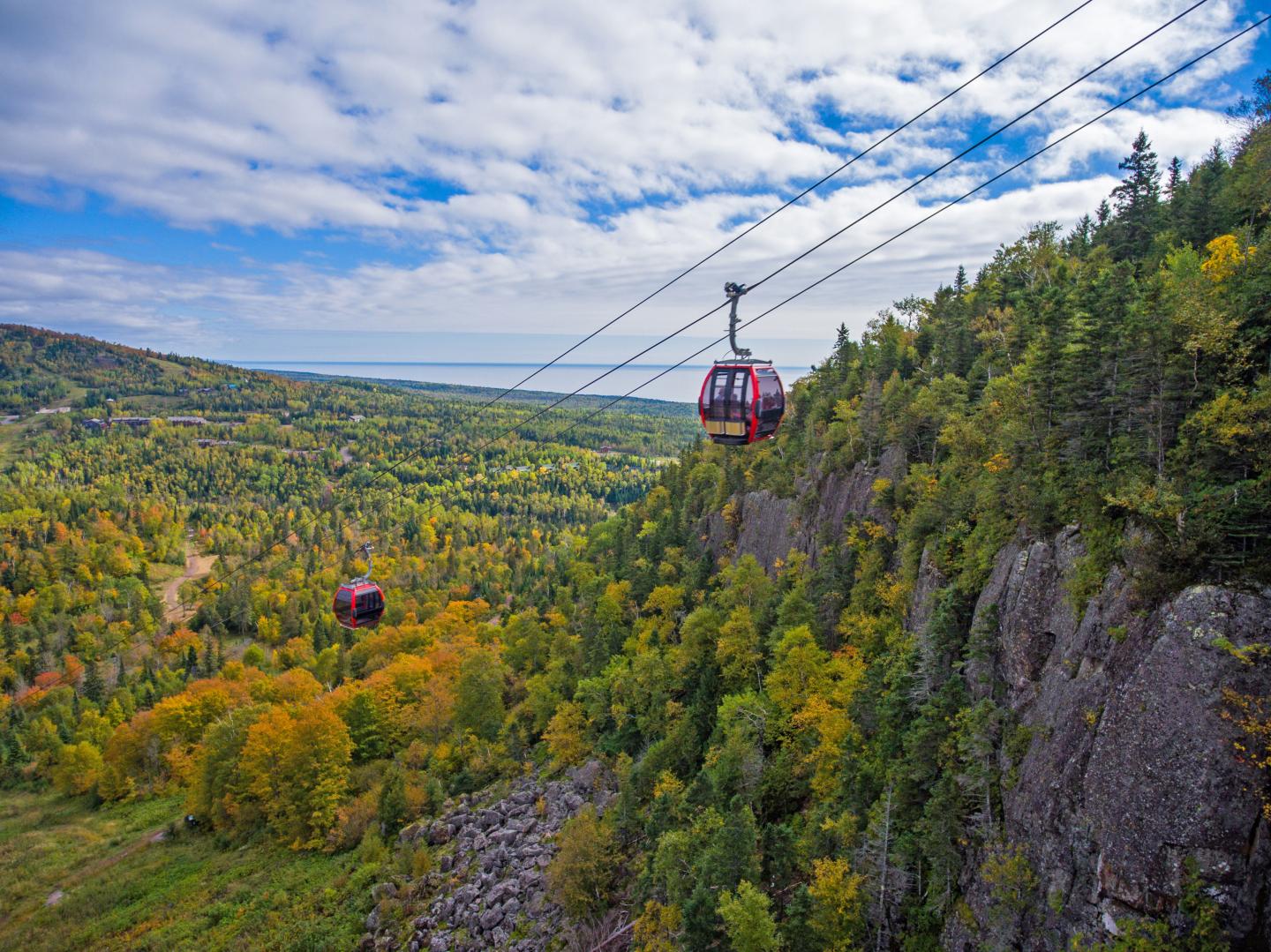 Cable cars over a scenic, forested valley with autumn foliage.