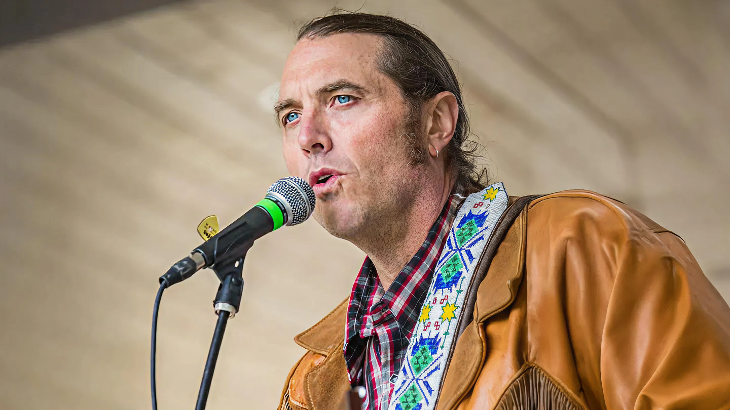 Singer at microphone wearing a brown jacket and colorful shirt.