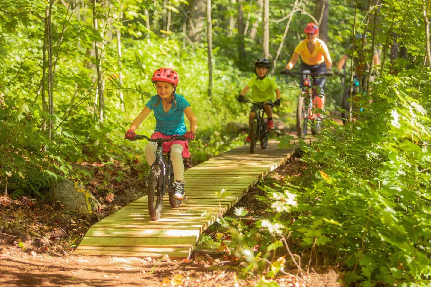 Children biking on a wooded trail bridge in bright clothing and helmets.