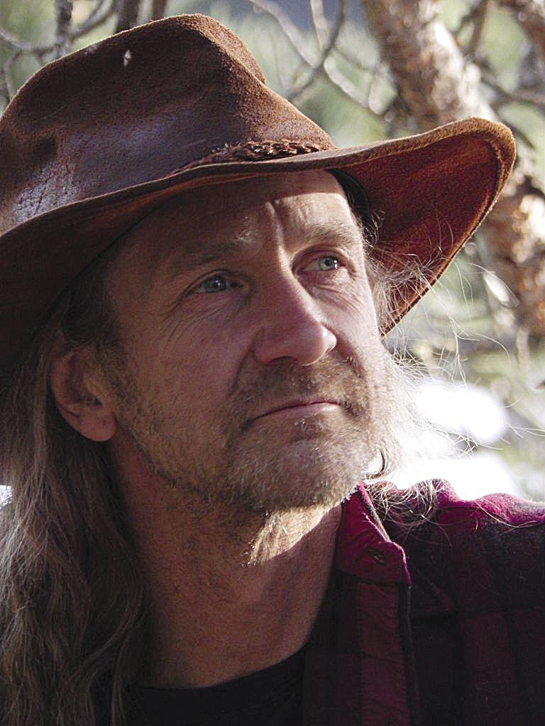 Man in a brown hat and flannel shirt, gazing thoughtfully outdoors.