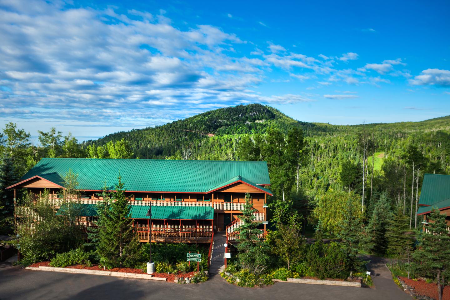 Lodge with a green roof surrounded by lush forest and hills under a blue sky.