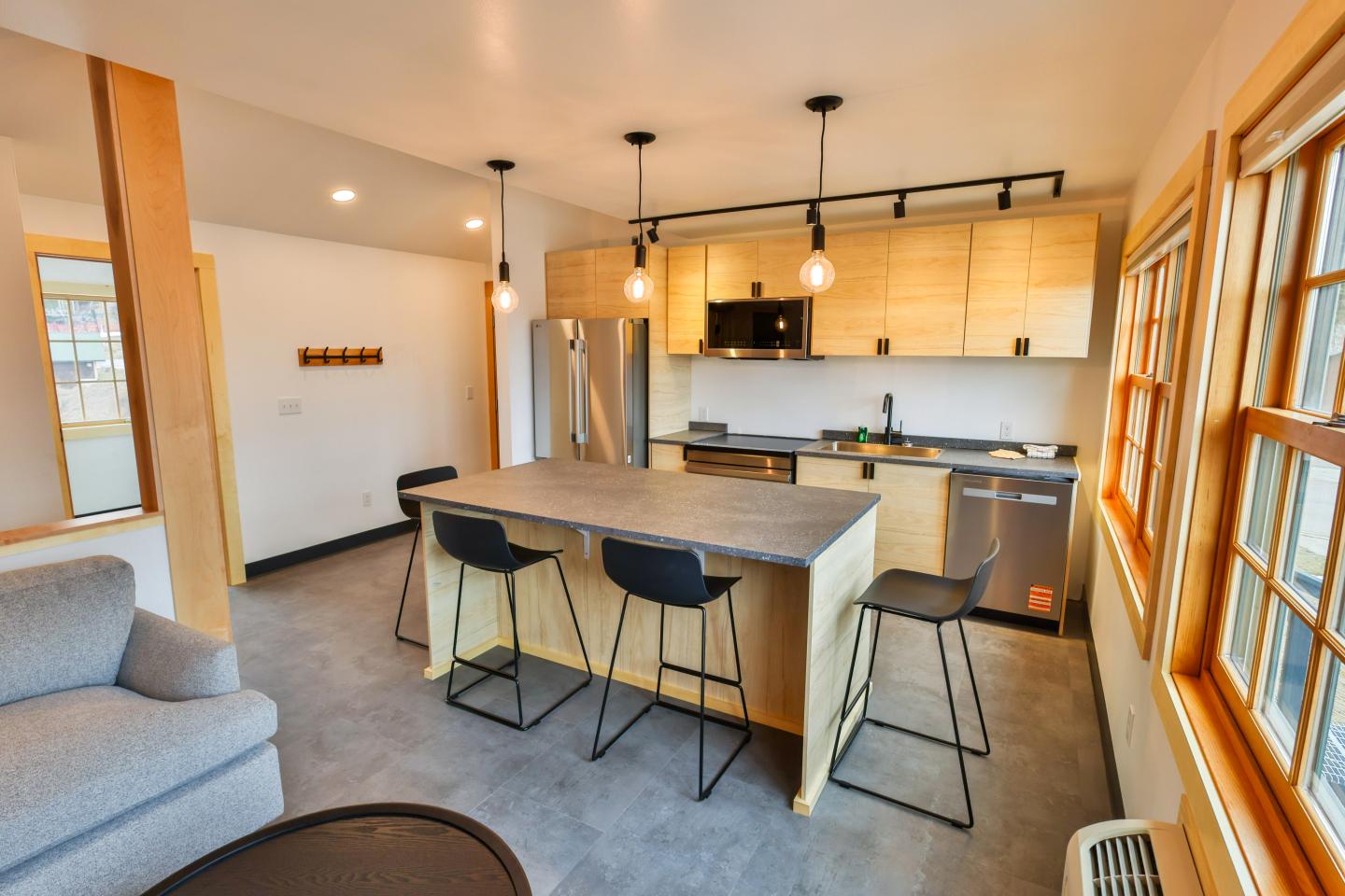 Modern kitchen with wooden cabinets, island, and black chairs. Bright lighting and large windows.