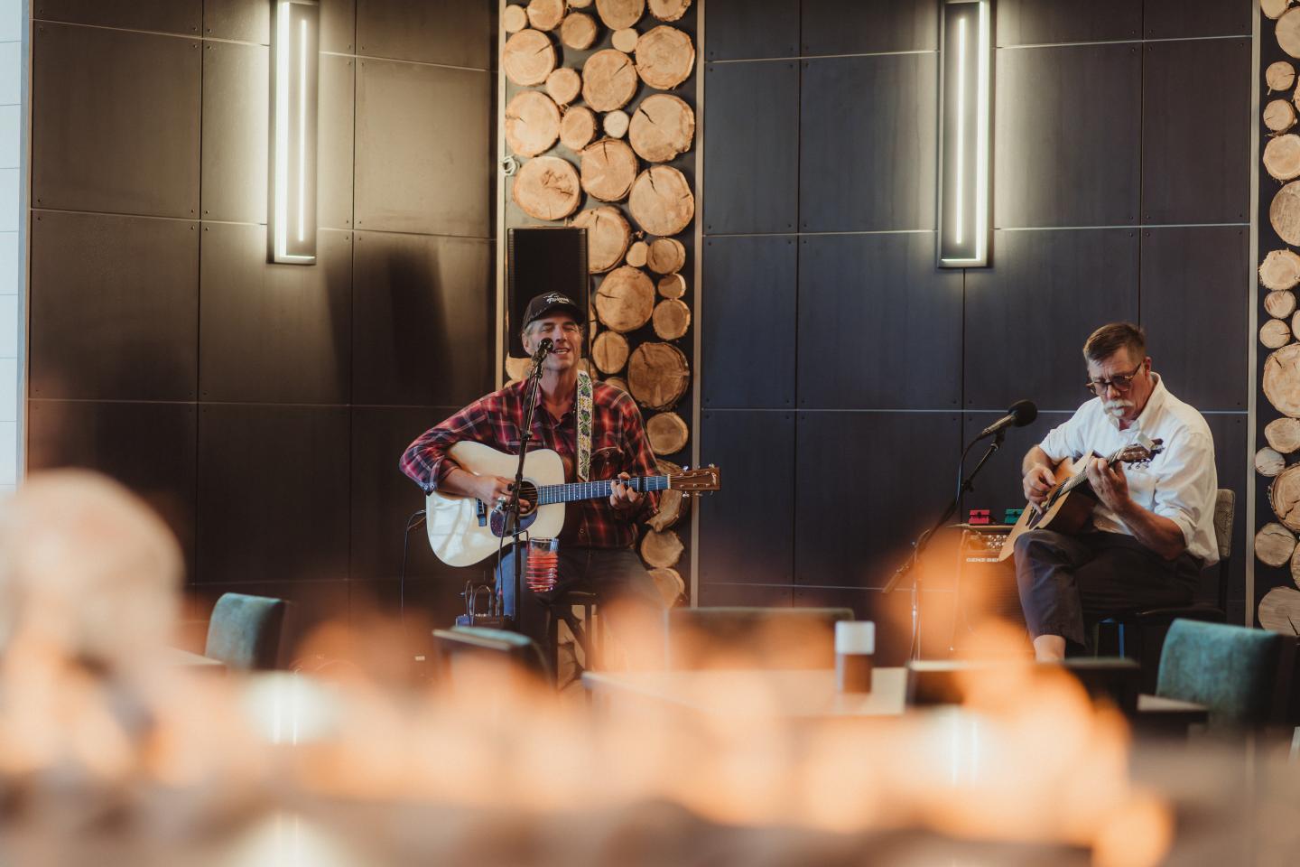 Two musicians playing guitars in a stylish, wood-accented room.