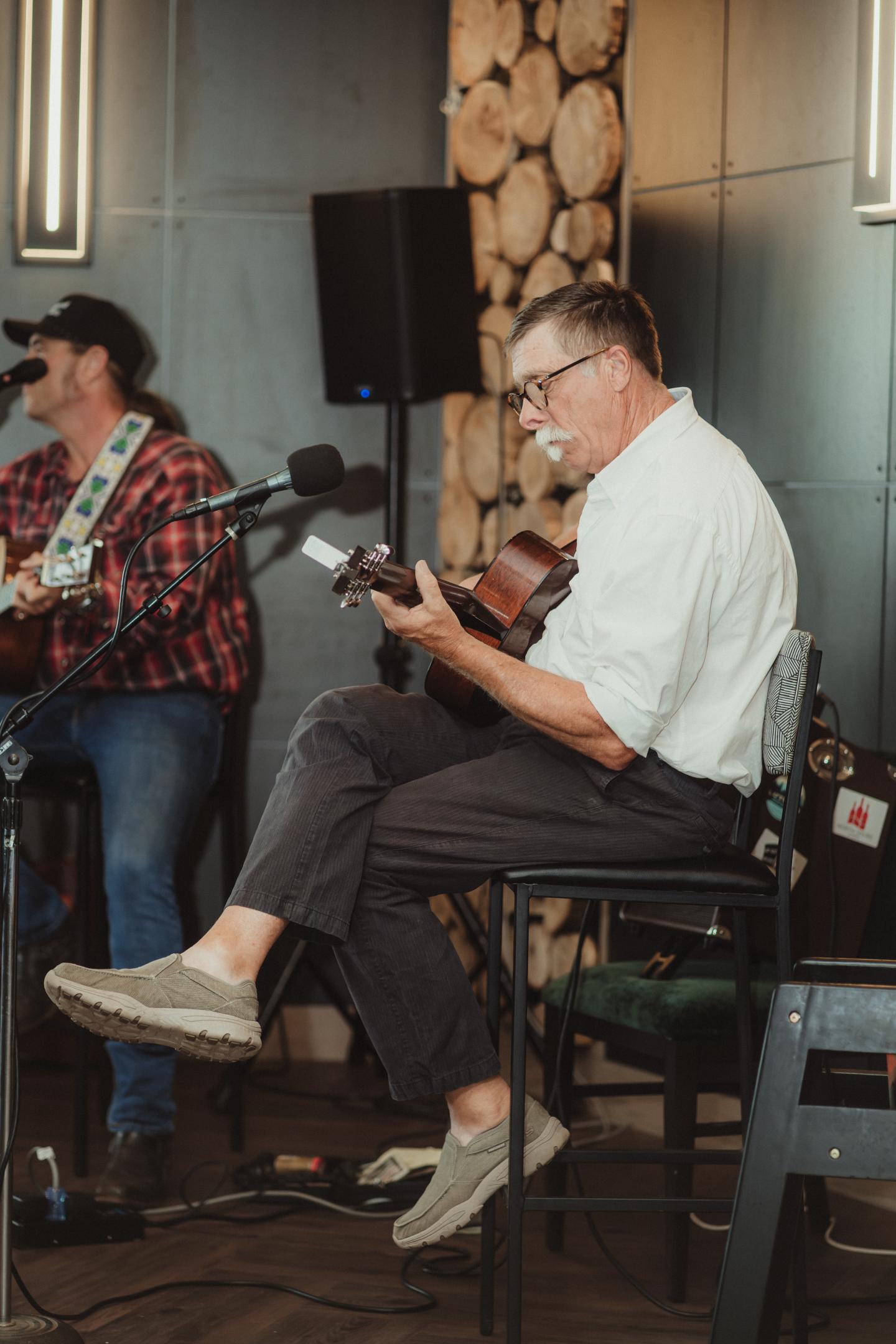 Man playing guitar on stage, wearing white shirt and glasses.