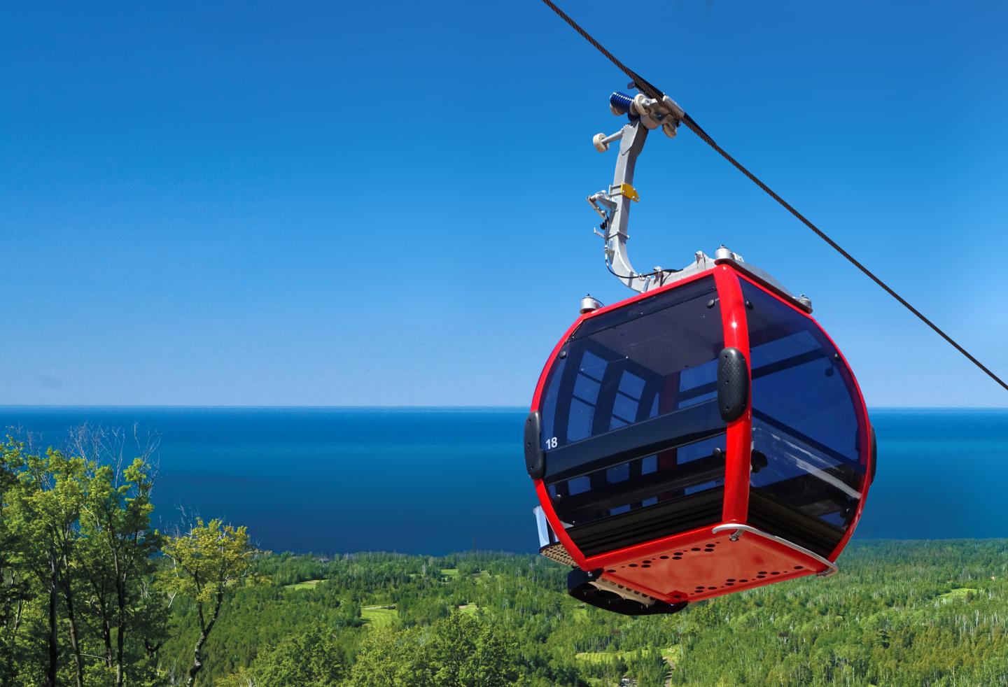 Red gondola over lush greenery near a blue ocean under clear sky.