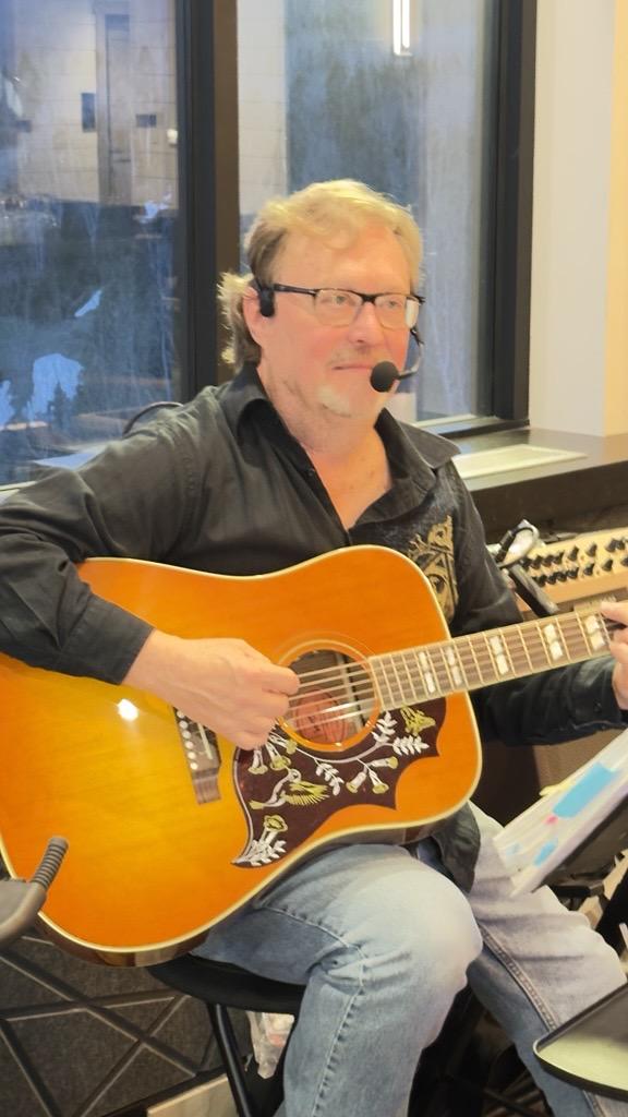 Man playing acoustic guitar with a headset in an indoor setting.