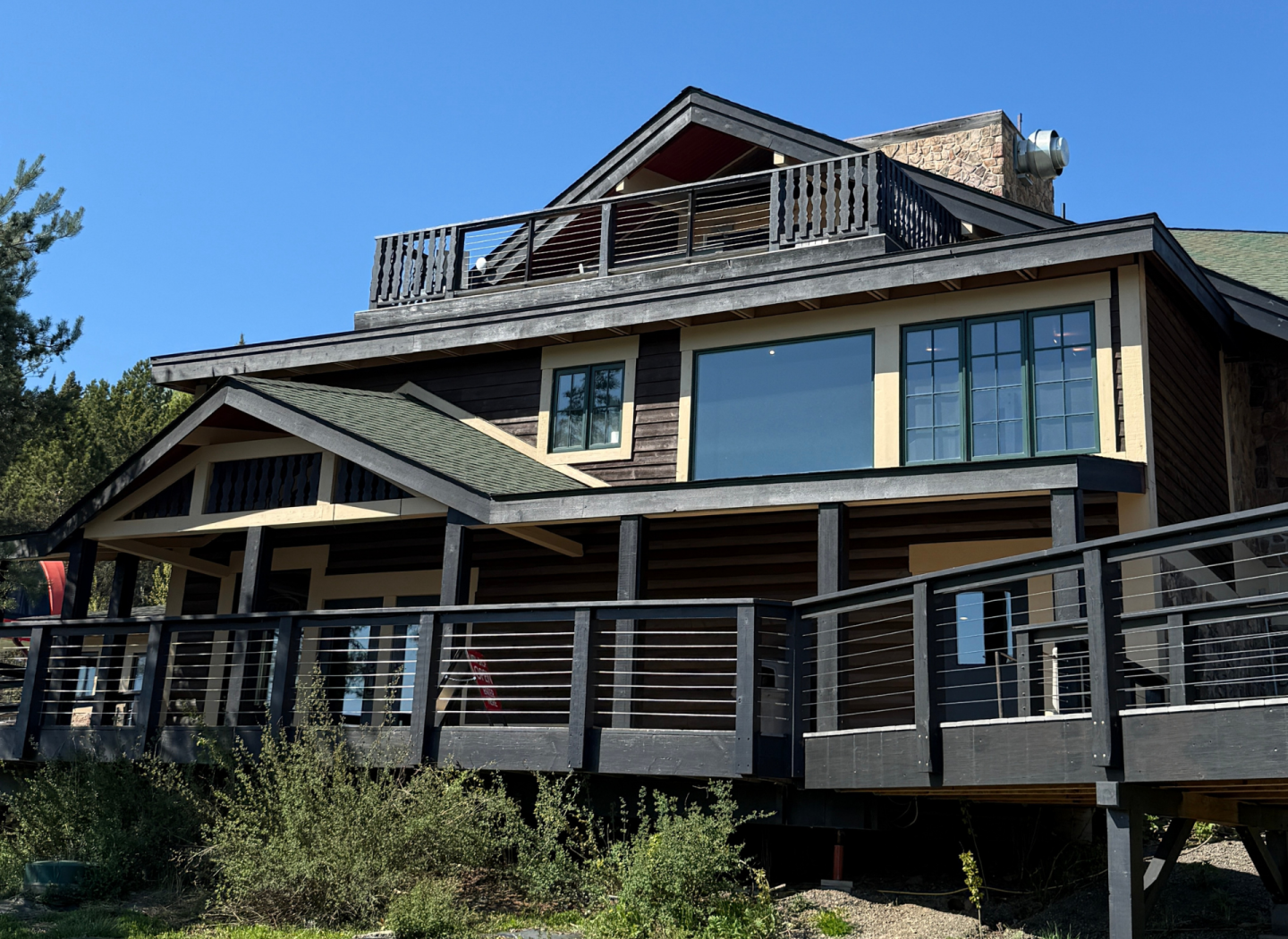 Large, rustic cabin with wooden deck, surrounded by greenery under a clear blue sky.