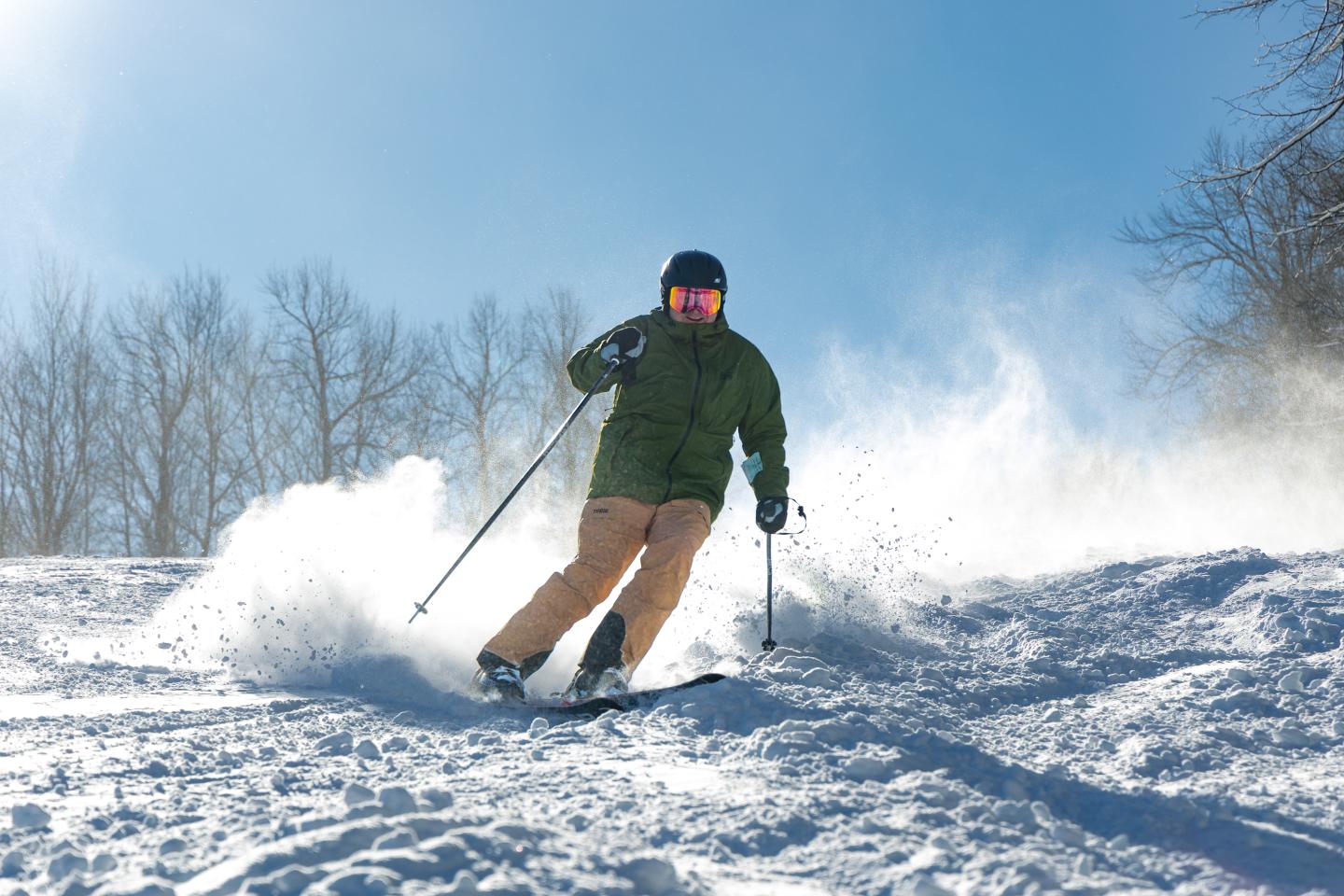 Skier in green jacket and tan pants descending snowy slope under clear blue sky.