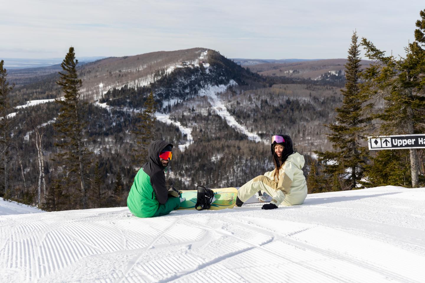 Two snowboarders resting on a snowy mountain slope, trees and hills in the background.