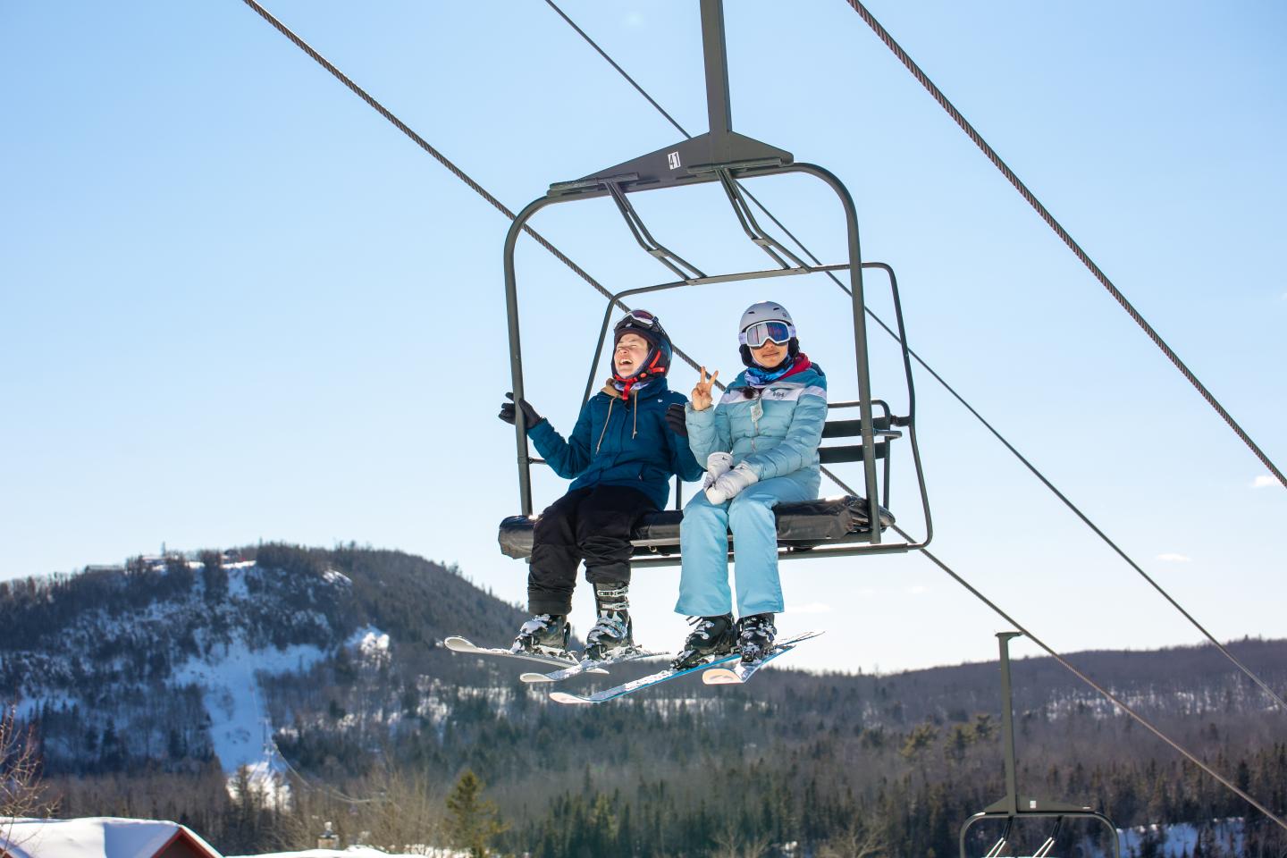Two people in winter clothing on a ski lift with snowy hills in the background.