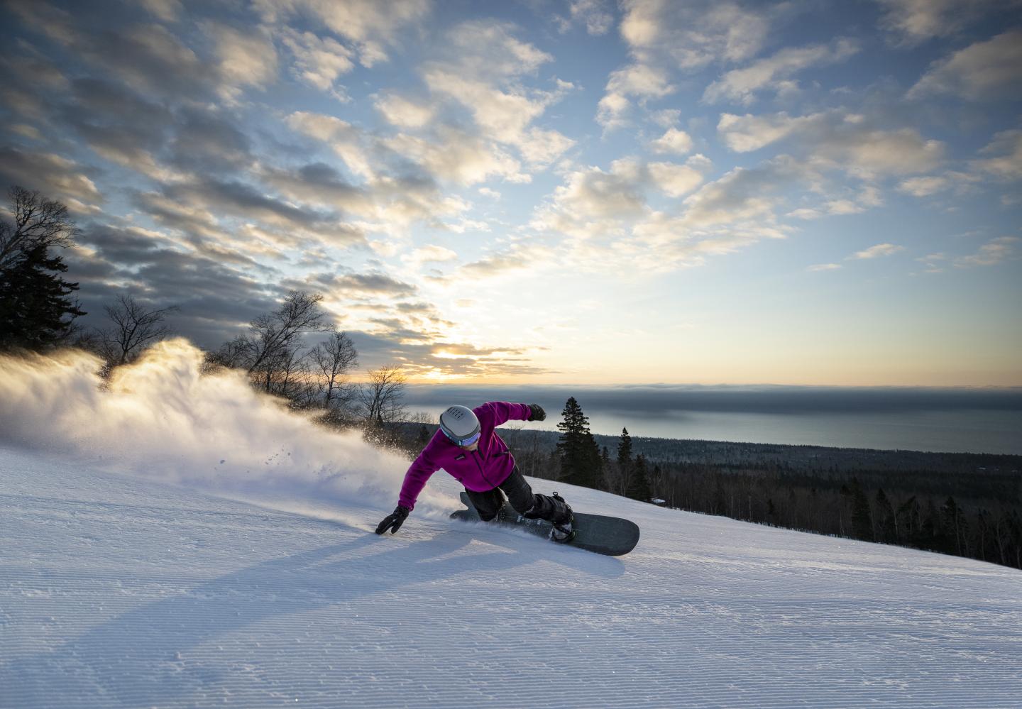 Snowboarder carving on a snowy slope at sunset, dramatic clouds above.