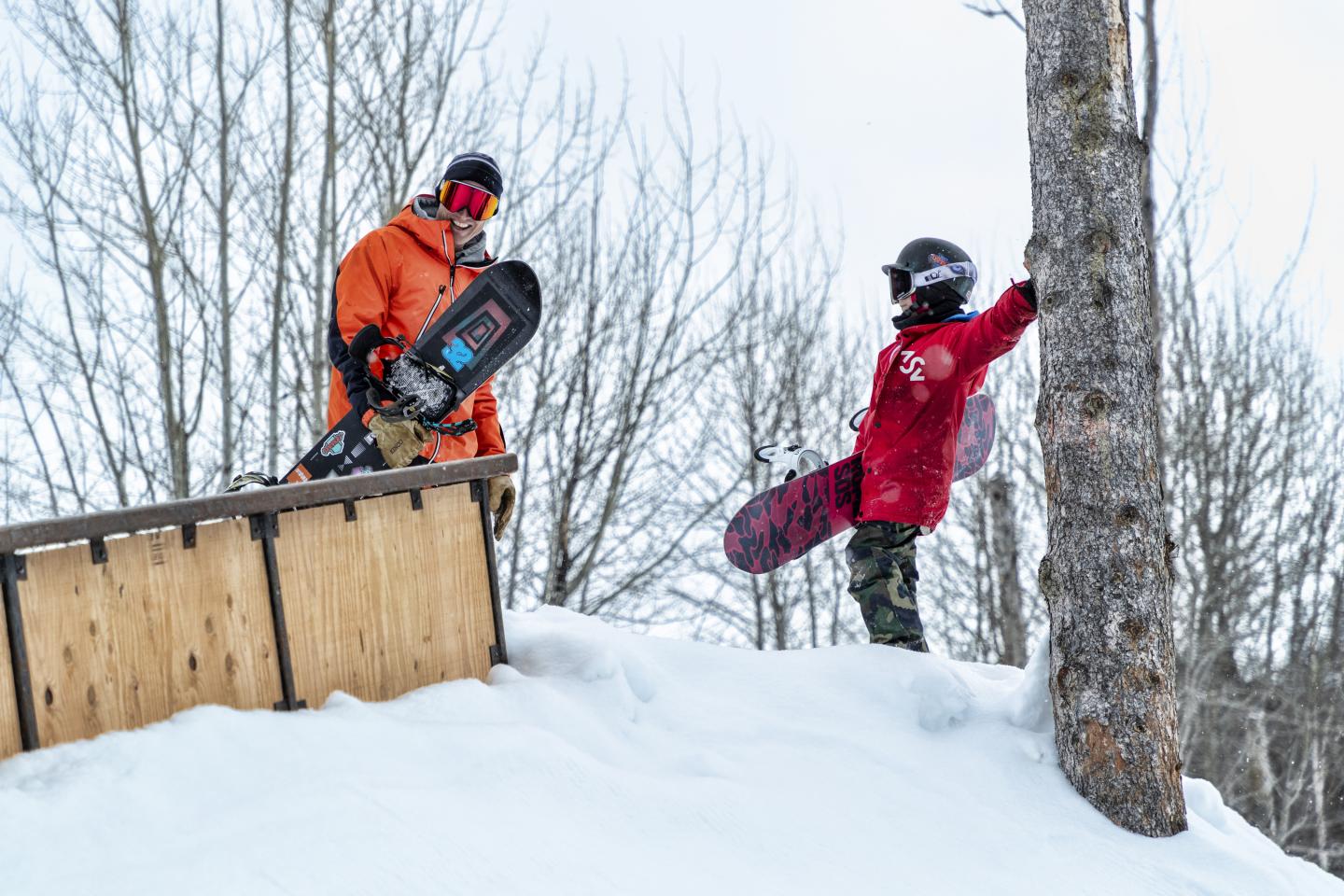 Two snowboarders in colorful jackets on a snowy hill by trees.