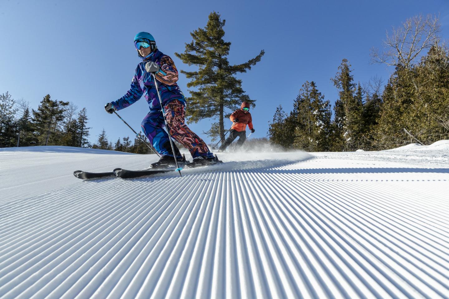 Two people skiing on a sunny, groomed slope with trees in the background.
