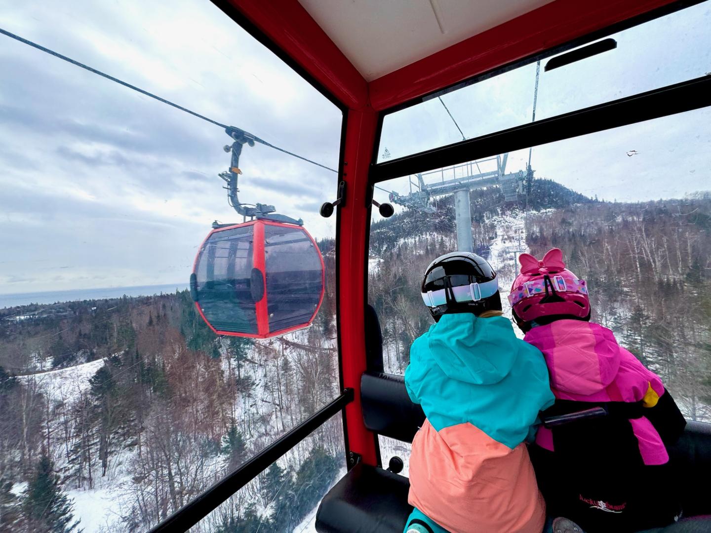 Children in ski gear riding a red gondola over a snowy landscape.