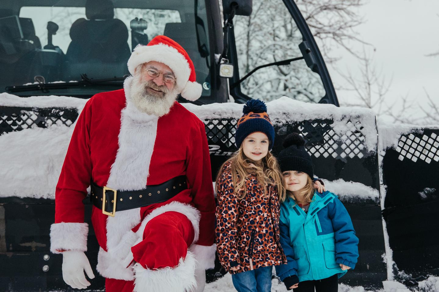 Santa Claus kneels beside two children in snowy winter clothing by a vehicle.