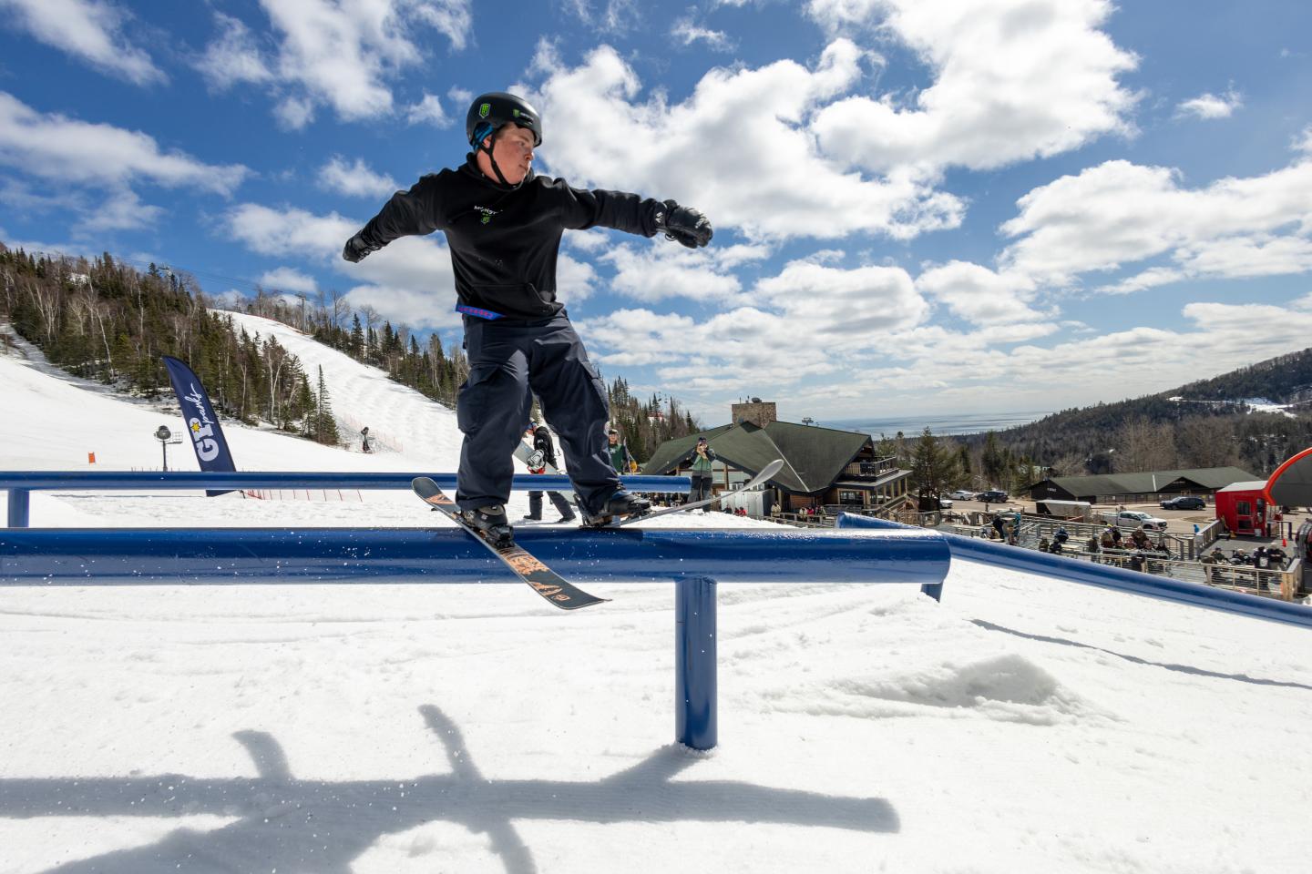 Skiier grinding a blue rail at a sunny mountain resort.