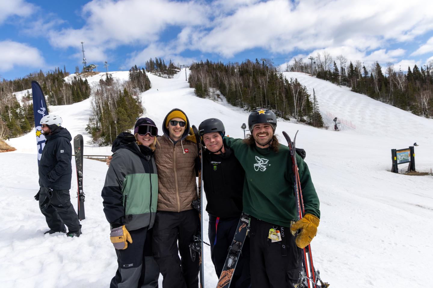 Four skiers in helmets and goggles pose together on a sunny snowy slope.