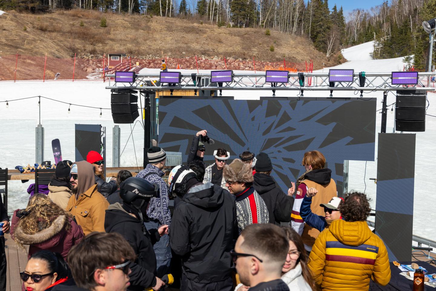 Crowd in winter clothing gathered at an outdoor music stage in snowy mountains.