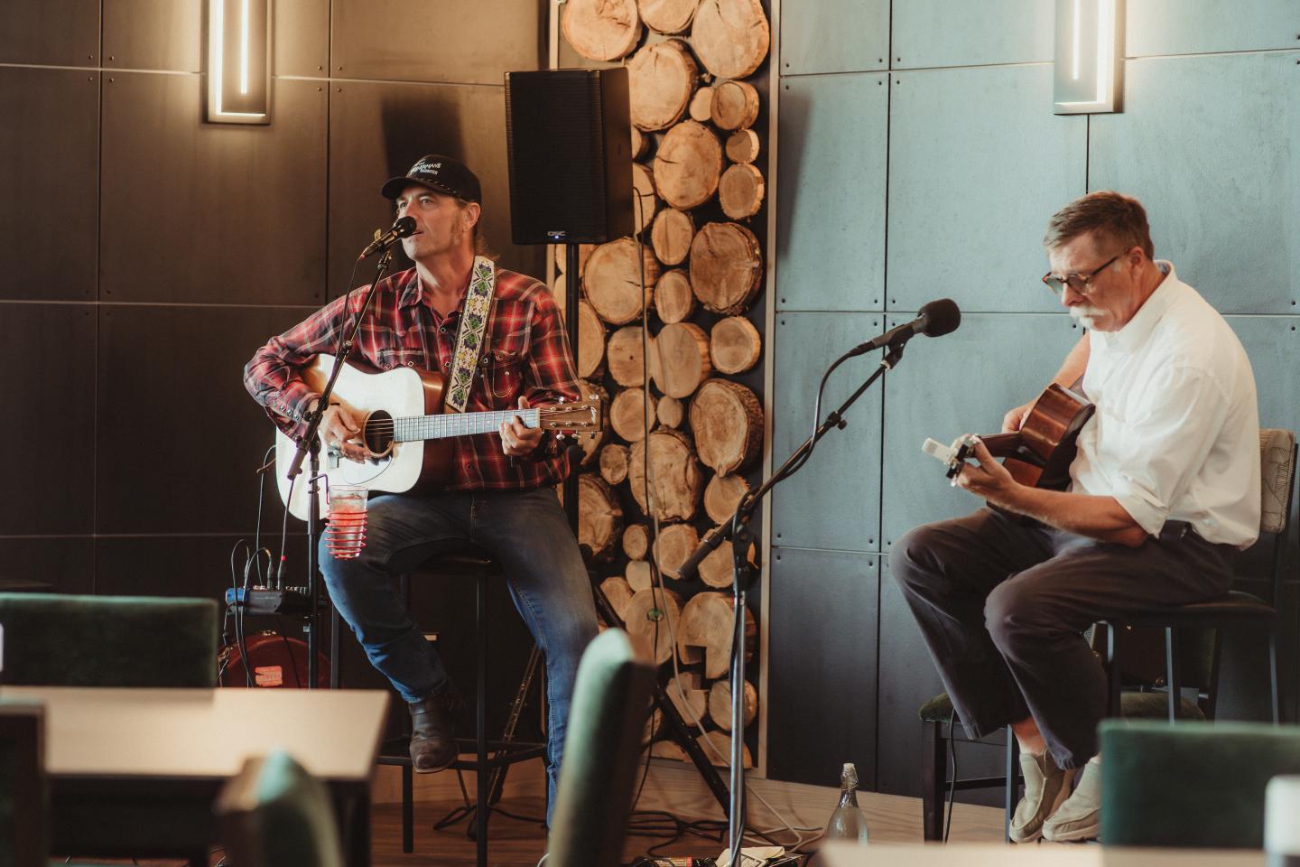 Two men playing acoustic guitars on a small stage beside stacked firewood.