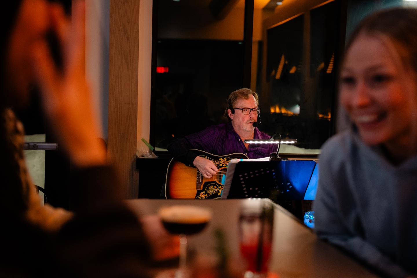 Man plays guitar in a warmly lit bar while a woman smiles in the foreground.